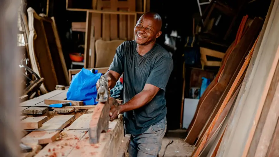 A smiling carpenter hand-planing a piece of timber in a workshop