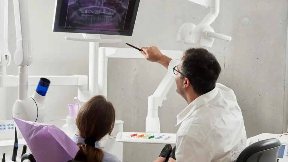 A dentist pointing at a dental X-ray on screen beside a patient