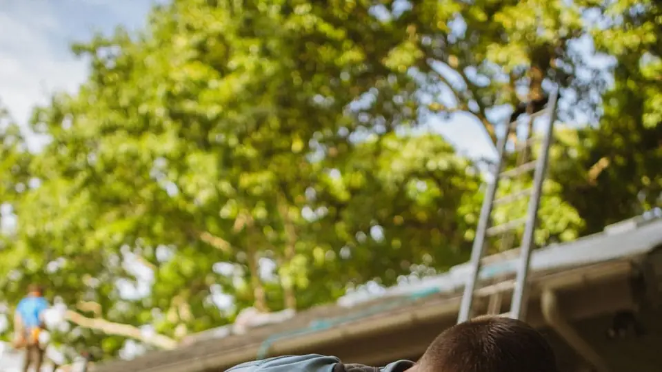 A roofer preparing tools with a team working on a roof behind